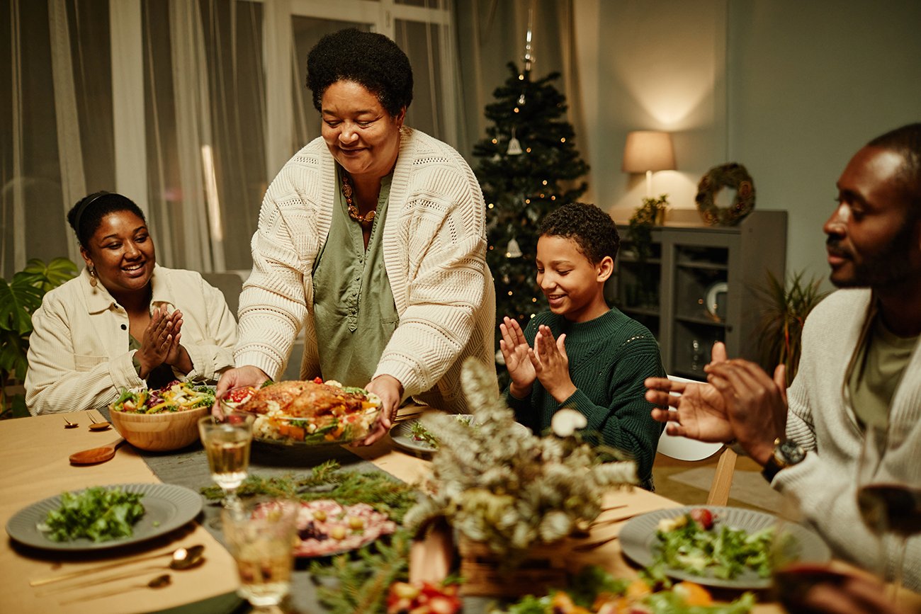 Family gathering around a festive table, enjoying a holiday meal with a woman serving a large dish, surrounded by cheerful family members, decorated space with a Christmas tree in the background.