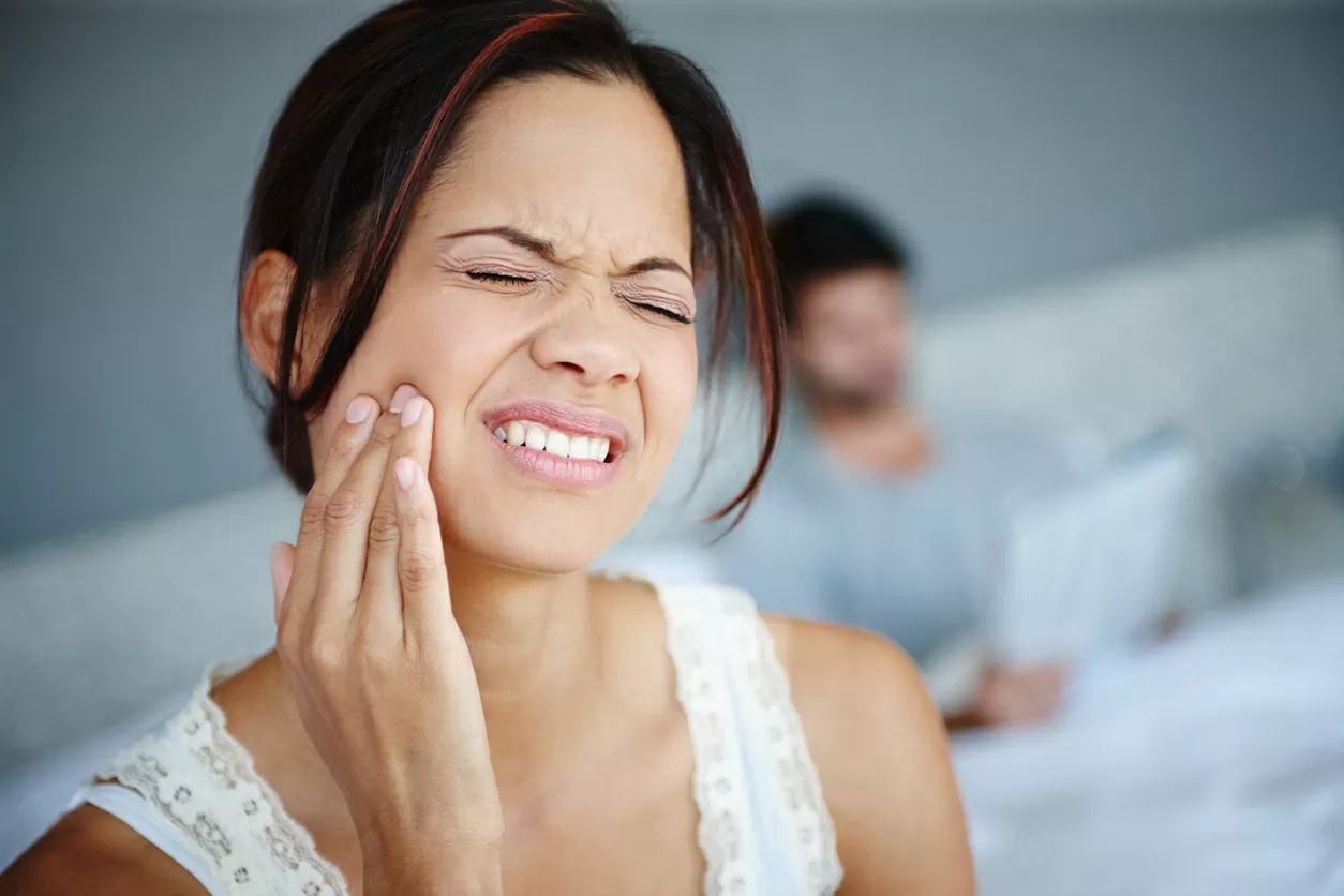 Woman experiencing jaw pain, indicating symptoms of TMJ disorder, with a blurred figure in the background.